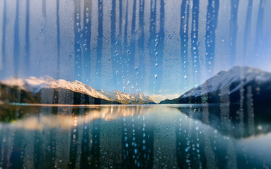 Poster - A view of a snow-capped mountain range reflected in a still lake through a misty window pane.