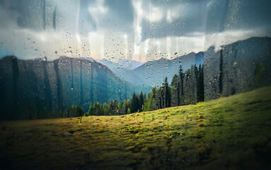 Poster - A view of a mountain range through a rain-streaked window. The sun peeks through the clouds, casting light on the green hills and dark trees.