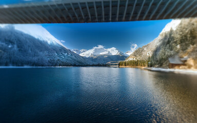 Poster - A scenic view of a mountain lake with snow-covered peaks and trees, seen through the underbelly of a bridge.
