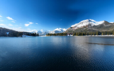 Poster - A panoramic view of a serene lake surrounded by snow-capped mountains, with a bright blue sky and a few puffy clouds.