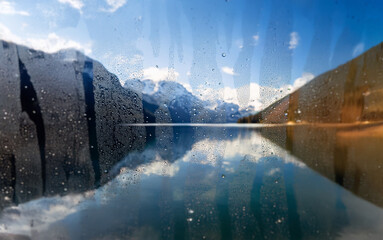 Poster - A view of a mountain lake through a wet window pane.  The reflection of the blue sky and snow-capped mountains can be seen in the still water.