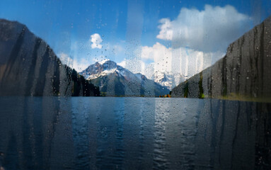 Poster - A view of a lake and snow-capped mountains through a rain-streaked window.