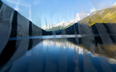 Poster - A view of snow-capped mountains reflected in a still lake, with a blurred foreground suggesting a windowpane.
