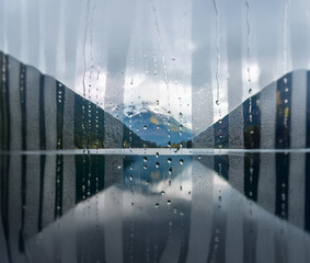Poster - A view of a mountain lake through a rain-streaked window, with a reflection of the cloudy sky and the mountains in the water.
