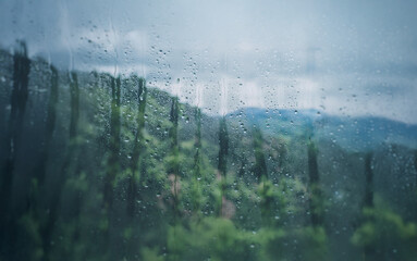 Poster - A raindrop-covered window reveals a lush, green landscape with rolling hills, the sky overcast with a soft, grey glow.