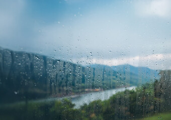 Poster - Raindrops on a windowpane obscure a view of a river, trees, and mountains.