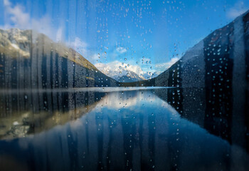 Poster - A view of a mountain range reflected in a still lake through a window covered in raindrops.
