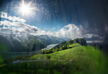 Poster - A view of a mountain range and a river, seen through a window with raindrops. The sun shines brightly behind the clouds.
