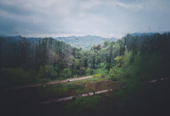 Aufkleber - A view of a forested valley through a rain-streaked window, with a blurred background of rolling hills and a cloudy sky.