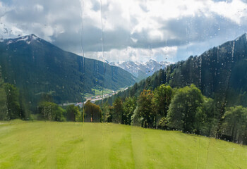 Poster - A picturesque view of a valley framed by mountains with a grassy field in the foreground, seen through a rain-streaked window.