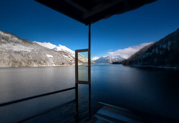 Poster - A view through an open door on a deck overlooking a still lake and snow-capped mountains on a clear day.