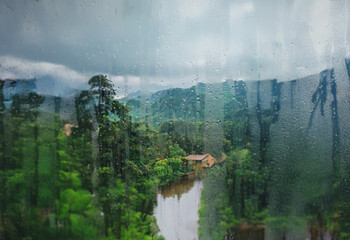 Poster - A view of a lush, green valley with a river and a small house through a rain-streaked window.