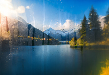 Poster - A view of a mountain lake through a wet windowpane.  The sun is setting, casting a warm glow on the scene.  The water is calm, and the mountains are reflected in the surface.
