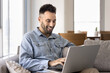 © fizkes - Happy Latin freelance businessman typing on laptop on home sofa, smiling, laughing, chatting online. Cheerful young man using Internet technology in pc, browsing social media on leisure time