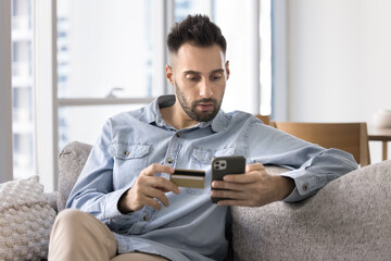 Serious Latin bank client man holding credit card, looking at mobile phone in shock, having financial problems with online account, finding mistakes in application work, lack of money