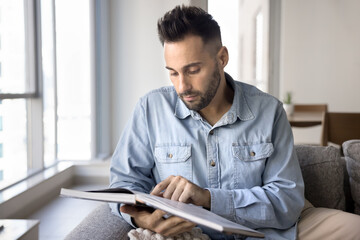 Focused handsome Latin man consulting data book, guide, keeping finger on page, reading textbook, studying at home, searching information for education. Freelance businessman checking paper notebook