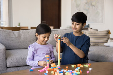 Two sweet Indian elder brother and younger sister kids stacking toy building blocks, playing in home living room, enjoying friendship, creative learning game, childhood, smart activity