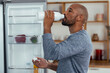 © Westend61 - Shaved head man drinking water from bottle near refrigerator at home