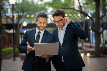 Two businessman on the street, deeply engrossed, eyes fixed on their laptop screen. Two handsome young businessmen in classic suits using laptop. Business men team using laptop outdoor. Business man