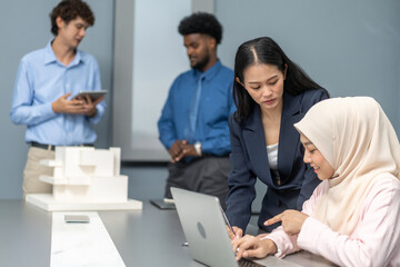 Muslim businesswoman in hijab discussing with asian colleague working on laptop in office, teamwork in a modern corporate environment, successful women collaborating on business projects, technology.