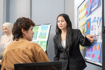 Young businesswoman leading a corporate presentation with global data charts and graphs, explaining business trends and strategies to colleagues during a team meeting in a modern office environment