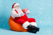 © deagreez - Full length photo of retired old man grey beard hold remote controll bucket watch sit orange beanbag wear red santa x-mas costume suspender 3d glasses cap boot isolated blue color background
