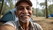 © Tom - Elderly South Asian man smiling warmly in a wooded campsite with tents in the background.
