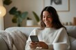 © Milos - A woman smiles as she looks at her smartphone, lounging on a cozy sofa wrapped in a knit blanket. The room is warmly lit and tastefully decorated.