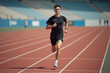 © Flash - fit young man with short hair wearing a black T shirt and dark pants running on a red track with a blurred stadium background