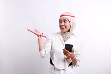 a happy asian muslim woman wearing red white headband, holding her phone, and pointing copy space be