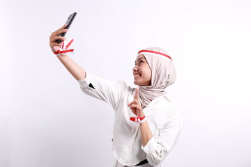 Happy young Asian muslim woman doing selfie shot on a cellphone isolated over white background. Celebrate Indonesian independence day on 17 August