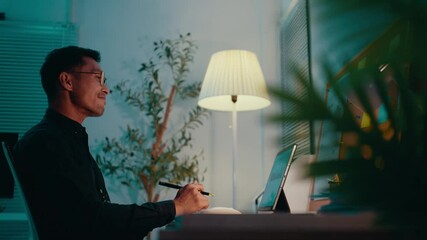 Poster - Young businessman working late in a home office, taking notes on a tablet while managing tasks at a desk. A modern workspace filled with technology and plants creates a productive atmosphere