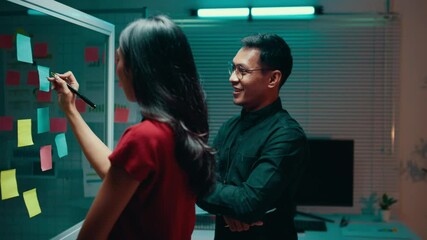 Poster - Asian businesswoman writes on sticky notes while discussing ideas with her partner in a nighttime office setting, showcasing collaboration and strategic planning