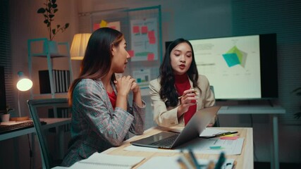 Wall Mural - Two young businesswomen engage in a discussion about a new project while working late at the office, showcasing collaboration and teamwork in a modern corporate environment