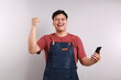 © ArvStd - Young asian man barista bartender barman employee in blue apron white t-shirt work in coffee shop using mobile cell phone do winner gesture isolated on white background. Small business startup concept