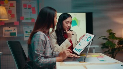 Poster - Two businesswomen are working late in the office, collaborating on a project. They are reviewing financial charts and data on a clipboard
