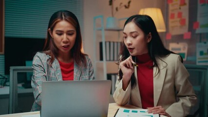 Poster - Two businesswomen are working late in their office. They are discussing a project and looking at a laptop. One of them is pointing at a document