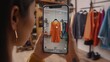 © Prostock-studio - A woman is holding her smartphone to view an orange dress in a virtual fitting room while surrounded by various clothing displays in a stylish boutique. The setting highlights technology and fashion.