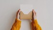© Elkhan Babayev - Female hands in yellow sweater hold an open craft paper envelope and a white card with blank space, taken from a high, first-person perspective on a white background.