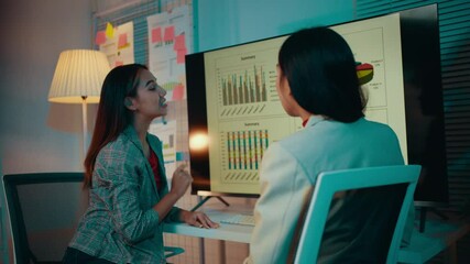 Poster - Two businesswomen are discussing positive results in their office at night. The woman standing is pointing at a computer screen with graphs