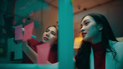 Poster - Two businesswomen having a discussion while using colorful sticky notes on a glass wall. They are working late at the office