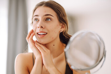 Wall Mural - Beautiful woman looking in the mirror, touching her face with her hand.  Pimples, acne, pores, scars. Natural skin without filters