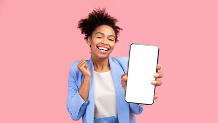 Poster - Happy black woman holding mobile phone with blank empty screen isolated over pastel pink wall, winner gesture. Mock up