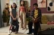 © pressmaster - Three preteens standing in comfortable living room, each holding their backpacks and school supplies, surrounded by cozy home decor and furniture