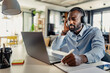 © lordn - African-American man wearing headphones, taking notes while working on a laptop from home, focused and engaged in a modern workspace.