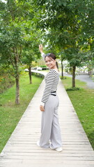 woman in striped shirt and gray pants poses joyfully on wooden pathway surrounded by greenery. Her cheerful expression adds to vibrant atmosphere of scene