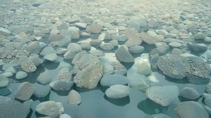 Fototapete - Water is slowly receding, revealing a riverbed with rocks of different sizes and shapes. The water reflects the sky, creating a surreal and peaceful atmosphere
