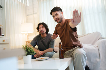 Happy Two Asian men watching sport games on television intently at sofa with focused expressions. Two friends enjoying football games. Friendship concept