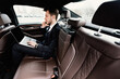 © Prostock-studio - Business Travel. Busy chief executive in suit using laptop, making phone call in luxury car, side view