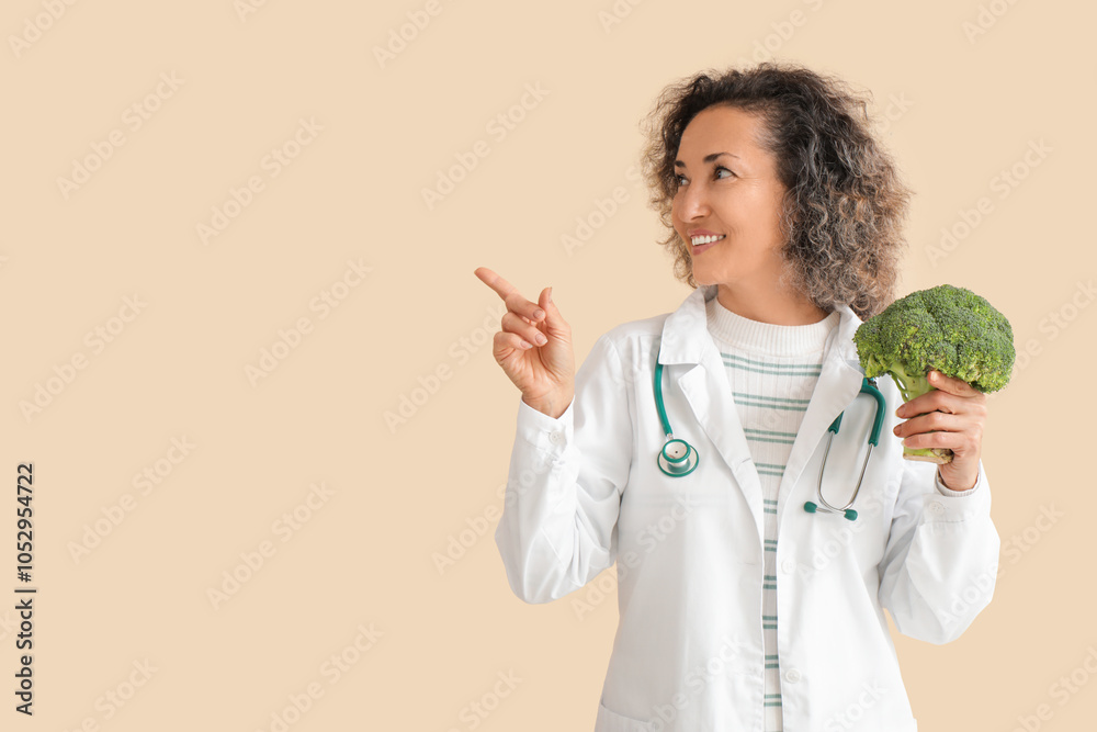 Mature female nutritionist with broccoli pointing at something on beige background